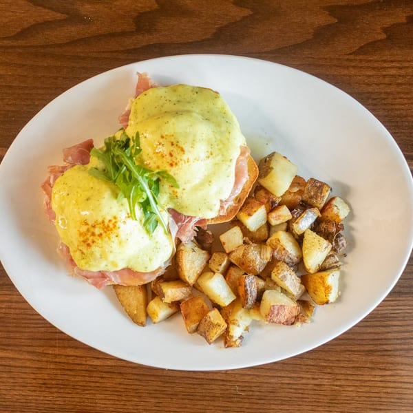 Eggs Benedict with hollandaise sauce and arugula, served with crispy home fries on a white plate.