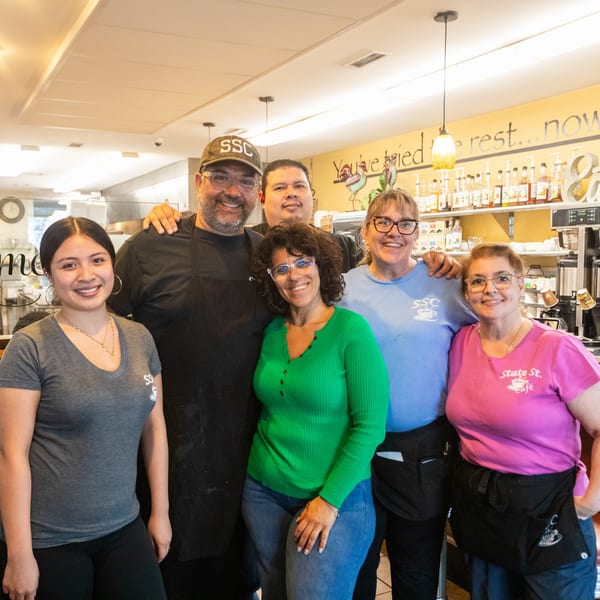 Six smiling State Street Cafe staff members, wearing branded shirts and aprons, pose together in the cafe.