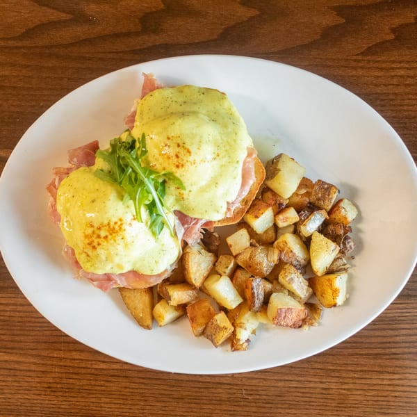 Eggs Benedict with prosciutto and arugula, served with roasted potatoes on a white plate.