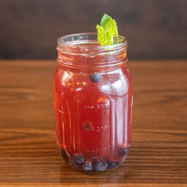 A mason jar filled with a red berry drink, garnished with mint and blueberries, on a wooden table.